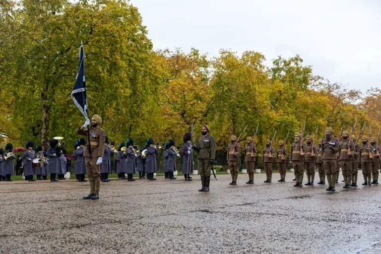 Soldiers in brown uniforms and turbans stand in formation during a ceremonial parade with a band in the background.