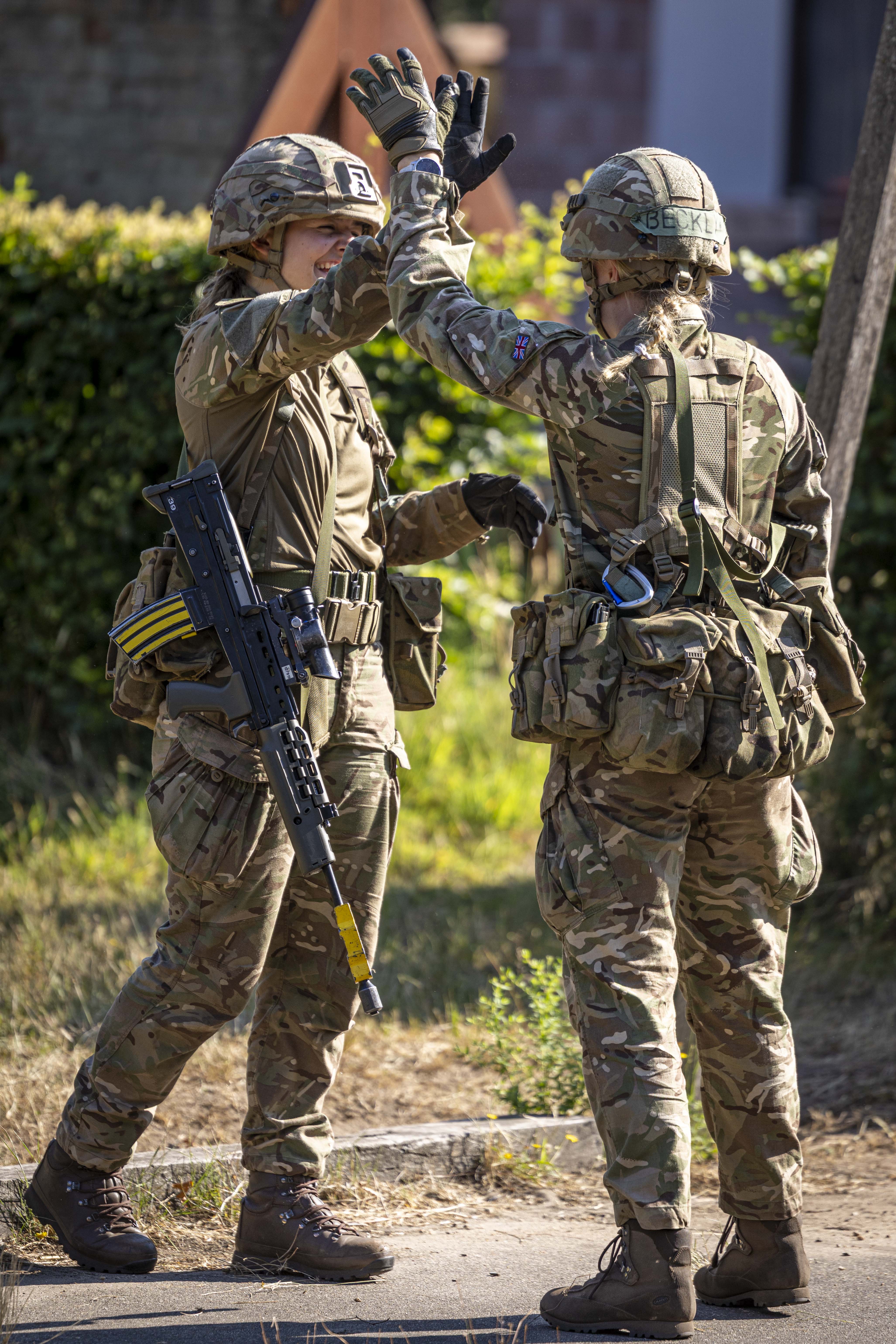 Two people in military uniforms high-five outdoors. One has a patch with 