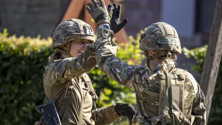 Two people in military uniforms high-five outdoors. One has a patch with