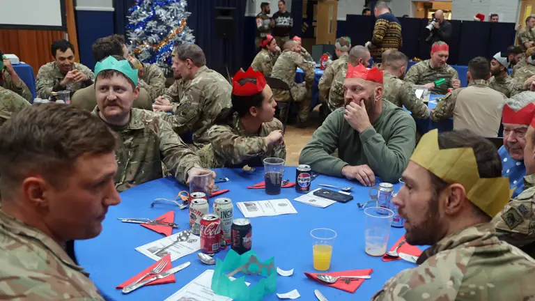 Soldiers wearing camouflage uniforms and Christmas hats sit around a table.