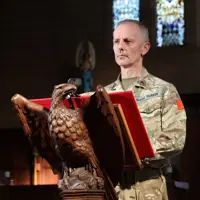 A person in military uniform stands at a carved eagle lectern in a church, appearing focused. Stained glass windows are visible in the background.