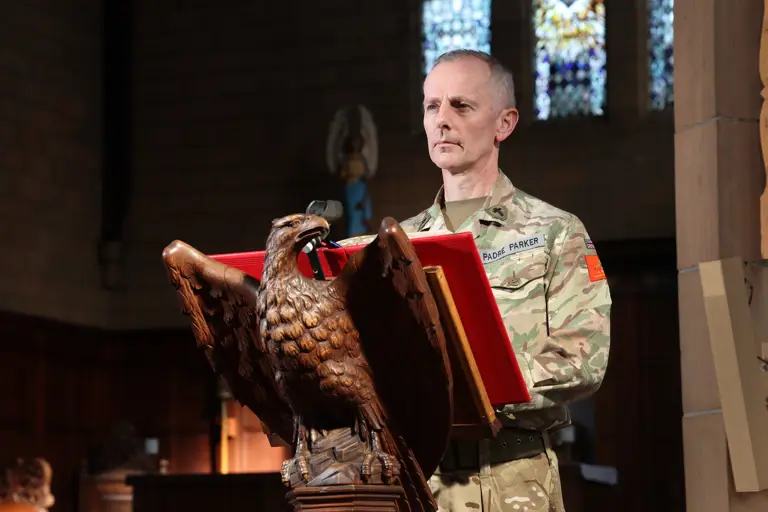 A person in military uniform stands at a carved eagle lectern in a church, appearing focused. Stained glass windows are visible in the background.