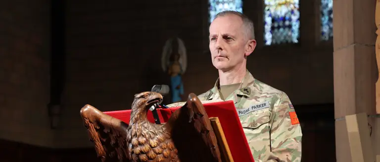 A person in military uniform stands at a carved eagle lectern in a church, appearing focused. Stained glass windows are visible in the background.