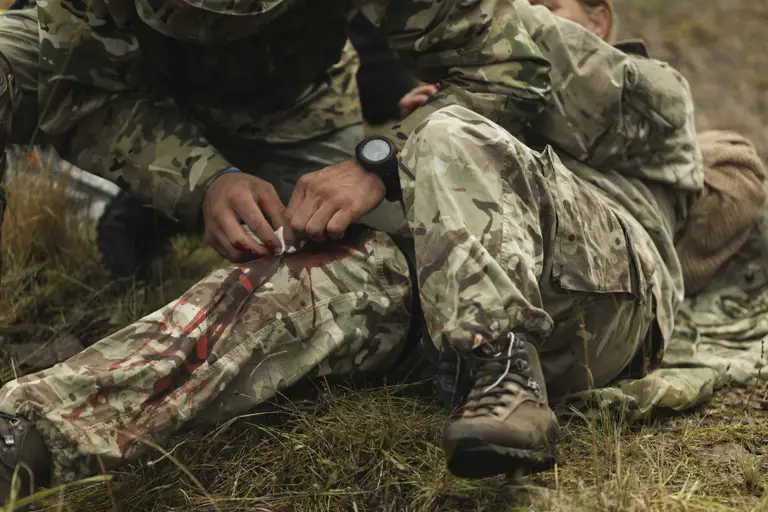 A soldier in camouflage tends to a bleeding leg wound of another on grassy terrain.