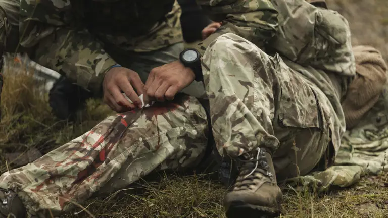 A soldier in camouflage tends to a bleeding leg wound of another on grassy terrain.
