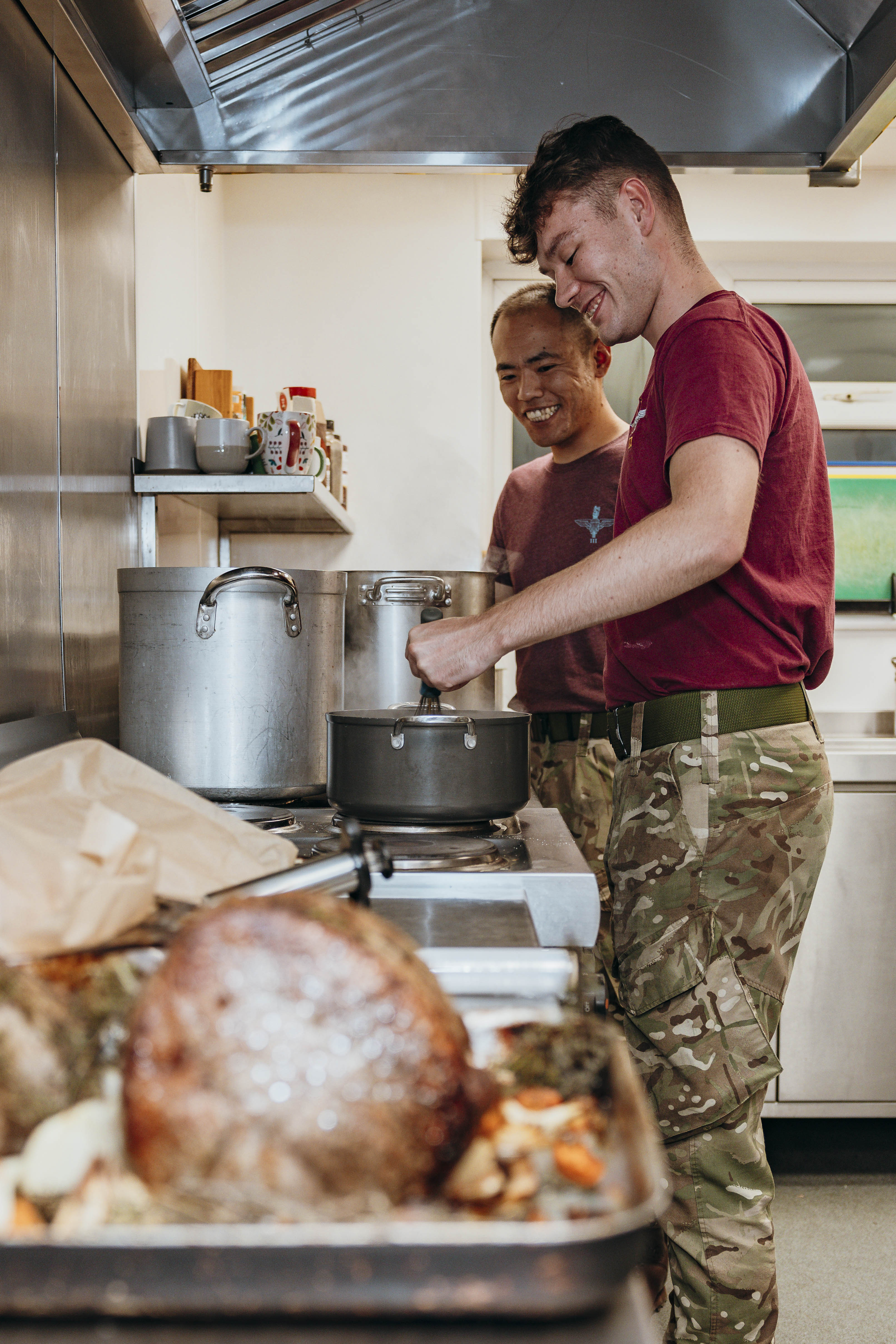 Two men in purple t-shirts and camo trousers smile as the man on the left whisks. 