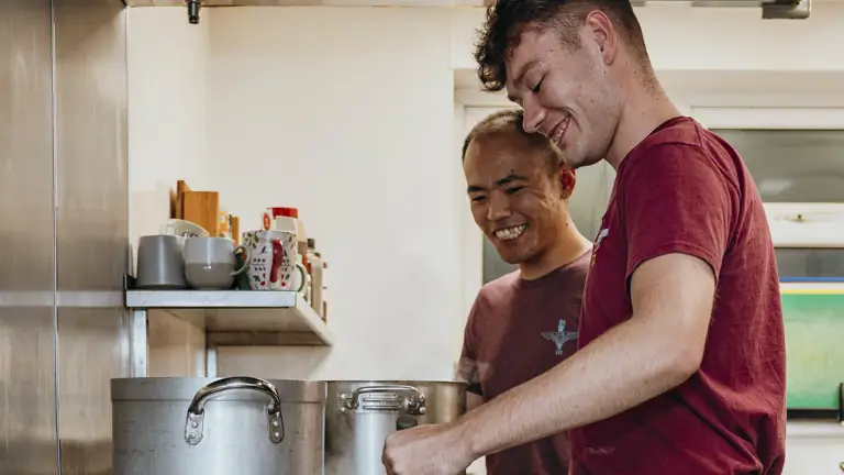 Two men in purple t-shirts and camo trousers smile as the man on the left whisks.