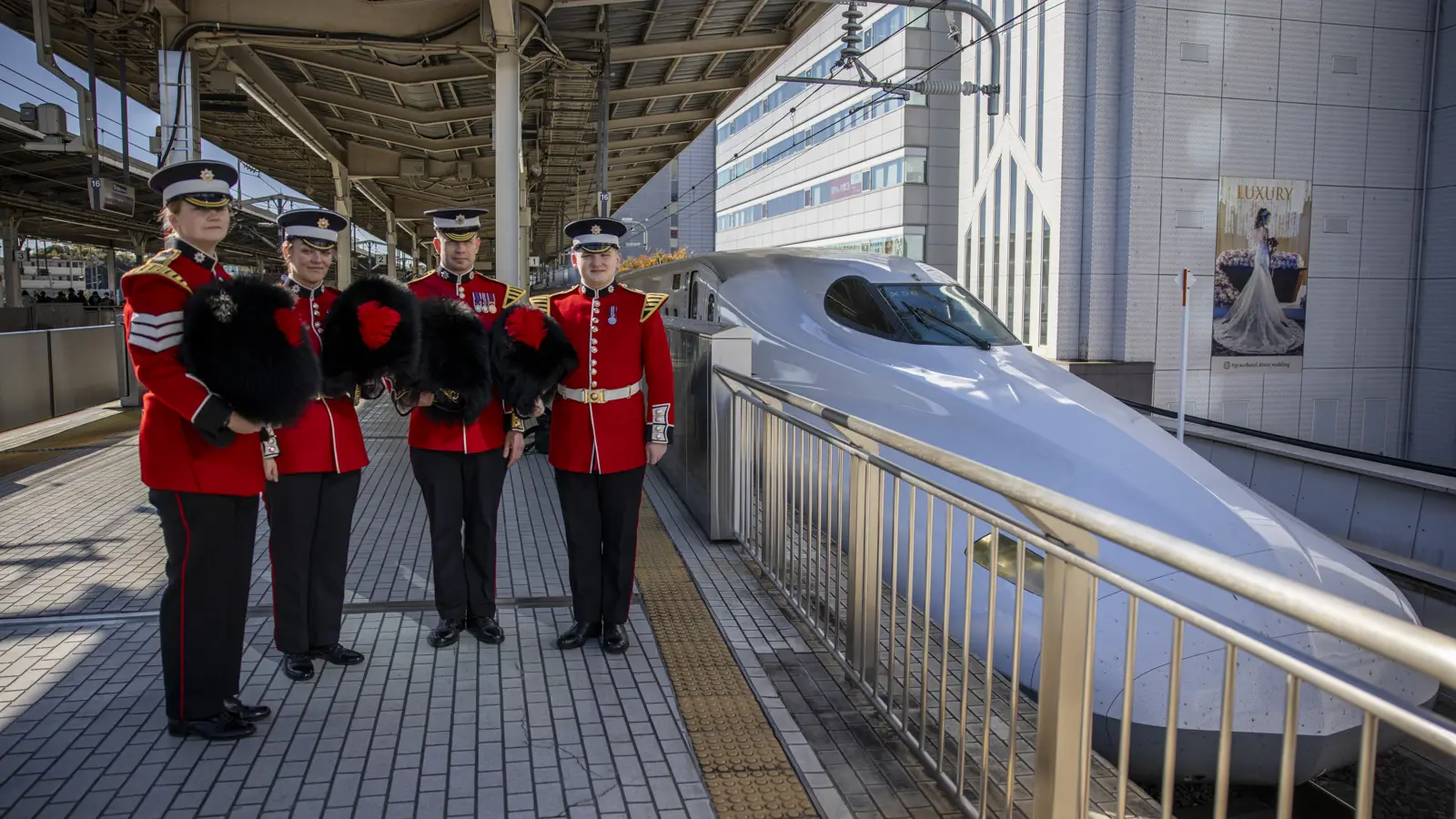 Four soldiers in red tunics and a bearskin hats are pictured next to a white bullet train.