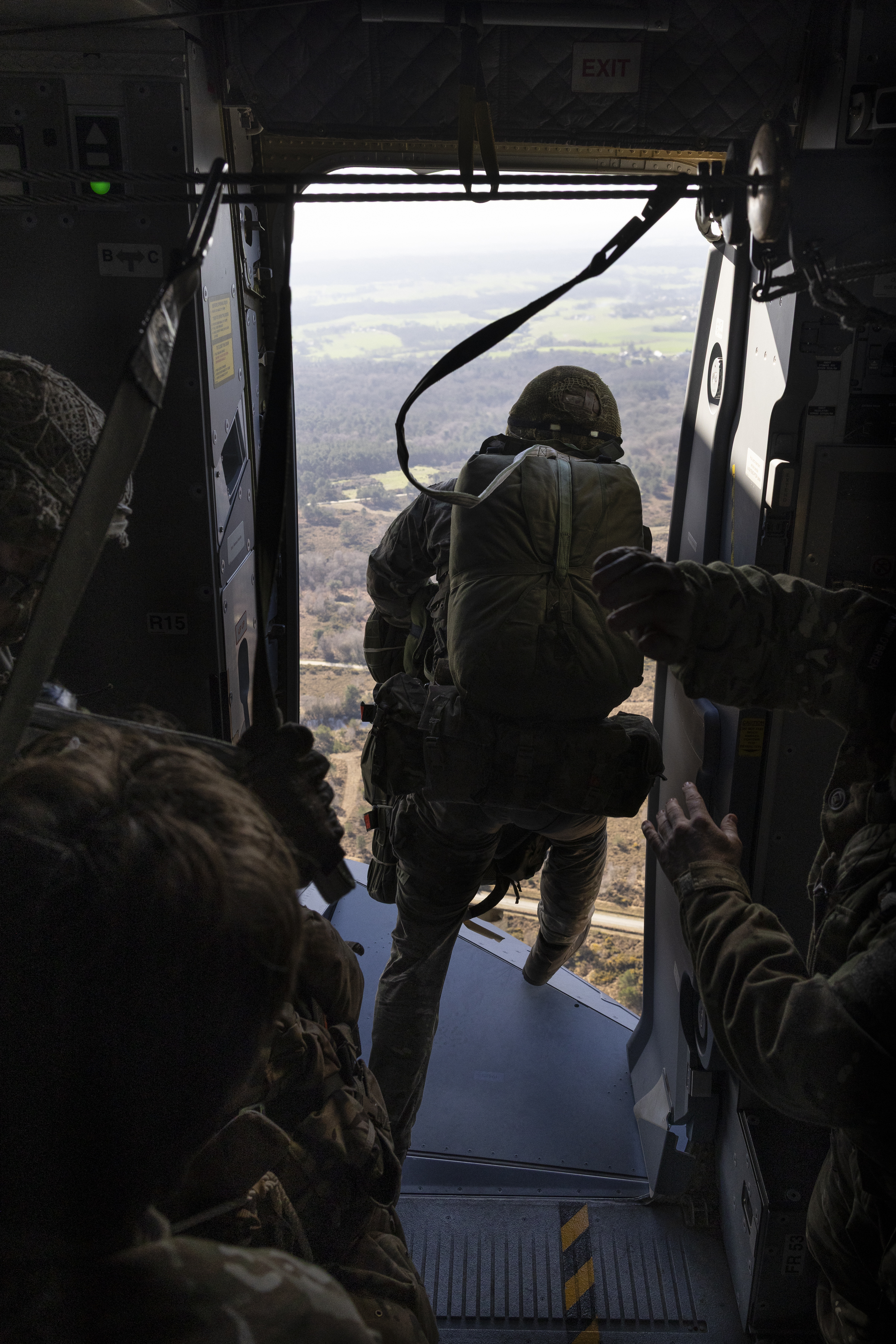 Soldier preparing to jump out of a military aircraft with a parachute harness over a rural landscape below.