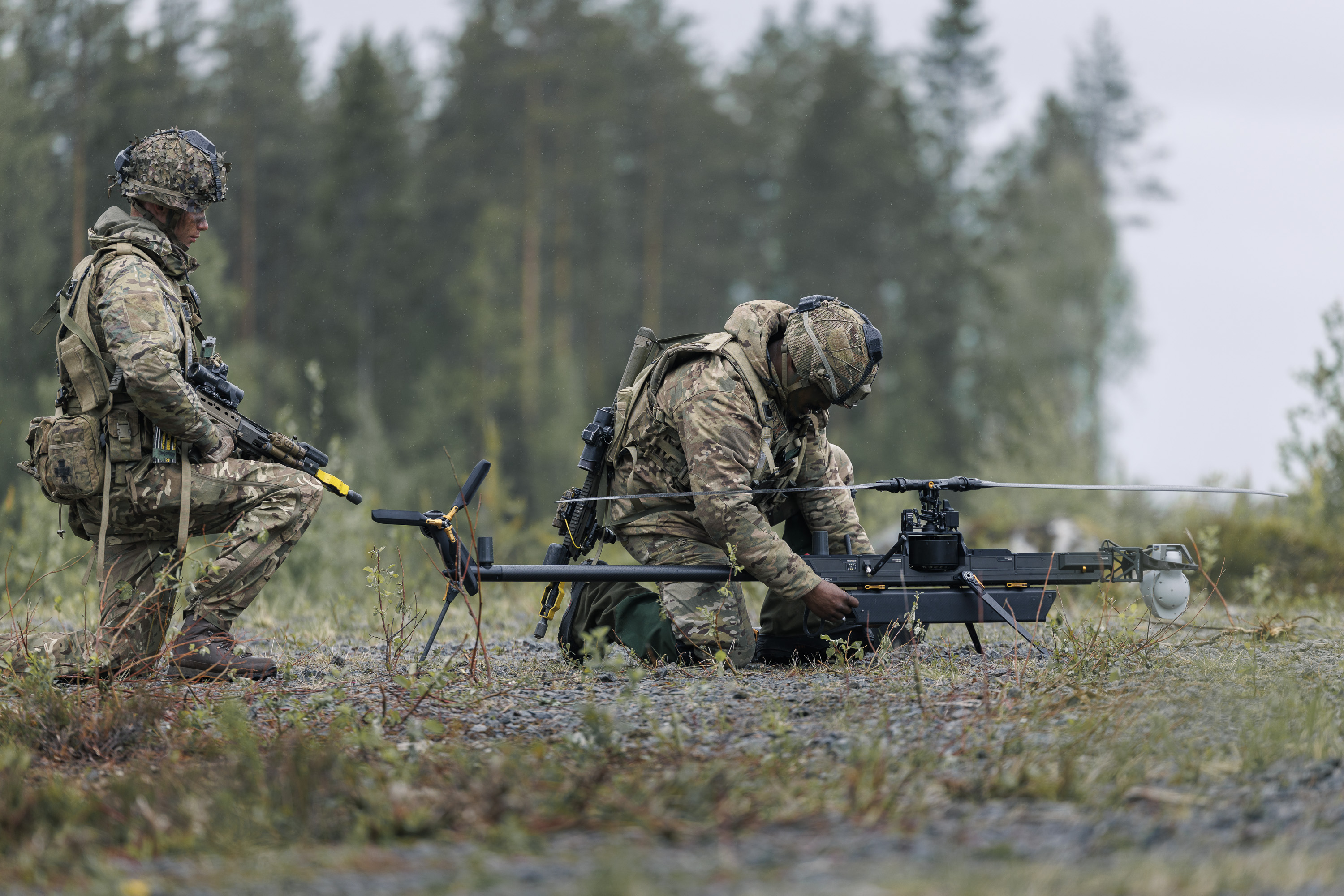 Two individuals in military gear are outdoors in a forested area, focusing on a small unmanned aircraft system on the ground. One is kneeling and adjusting the drone, while the other stands nearby holding equipment. The scene is set on a gravel surface with trees in the background.
