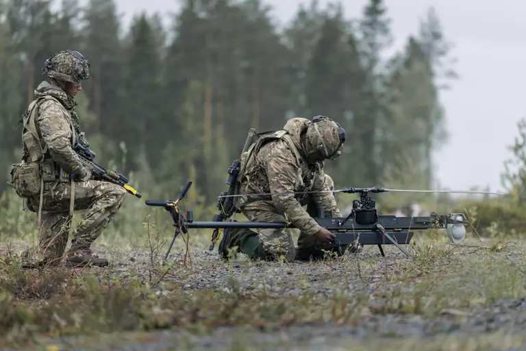 Two individuals in military gear are outdoors in a forested area, focusing on a small unmanned aircraft system on the ground. One is kneeling and adjusting the drone, while the other stands nearby holding equipment. The scene is set on a gravel surface with trees in the background.