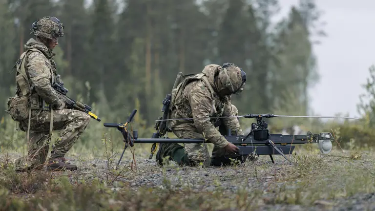 Two individuals in military gear are outdoors in a forested area, focusing on a small unmanned aircraft system on the ground. One is kneeling and adjusting the drone, while the other stands nearby holding equipment. The scene is set on a gravel surface with trees in the background.
