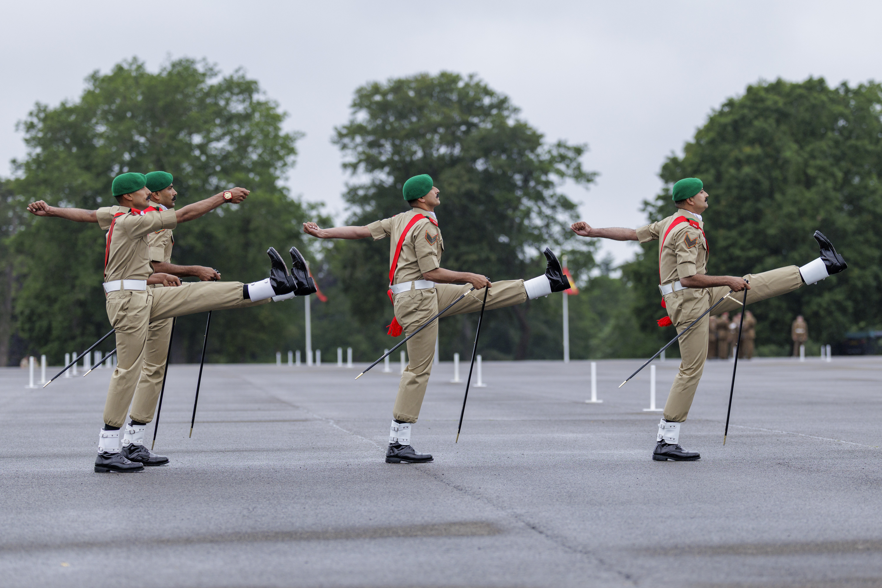 Four individuals in uniform, wearing green berets and red sashes, are performing a synchronized march with high kicks. They are on a wide, paved area with large trees in the background, possibly at a military or ceremonial event.
