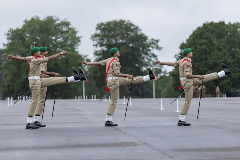 Four individuals in uniform, wearing green berets and red sashes, are performing a synchronized march with high kicks. They are on a wide, paved area with large trees in the background, possibly at a military or ceremonial event.