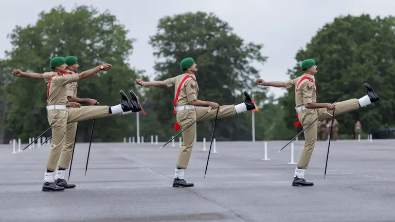 Four individuals in uniform, wearing green berets and red sashes, are performing a synchronized march with high kicks. They are on a wide, paved area with large trees in the background, possibly at a military or ceremonial event.