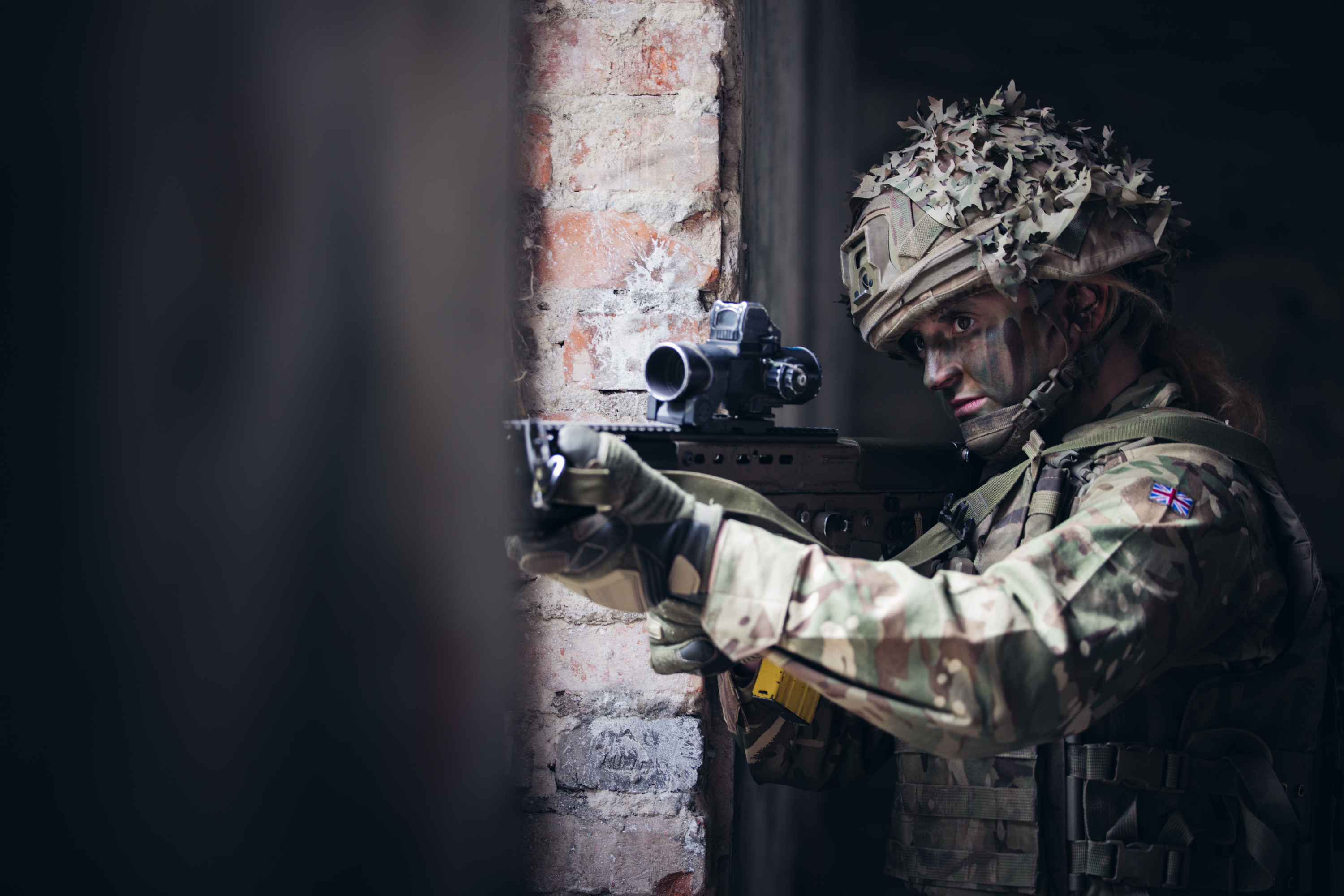Soldier in camouflage gear aiming a rifle with a scope inside a dimly lit brick building.