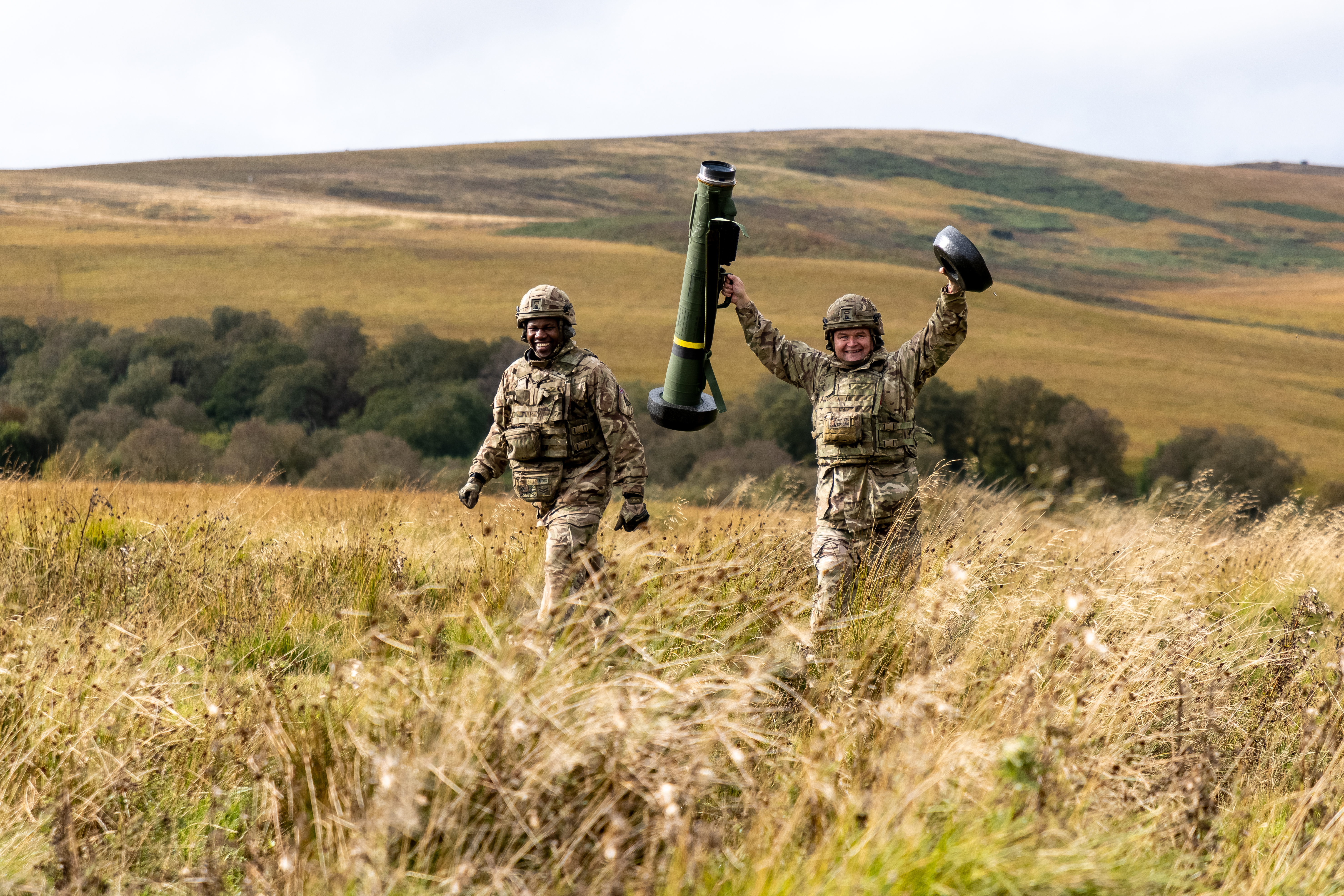 Two soldiers in uniform smile and one holds a missile launcher in the air. 