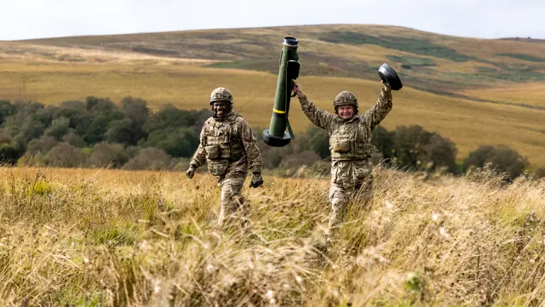 Two soldiers in uniform smile and one holds a missile launcher in the air.