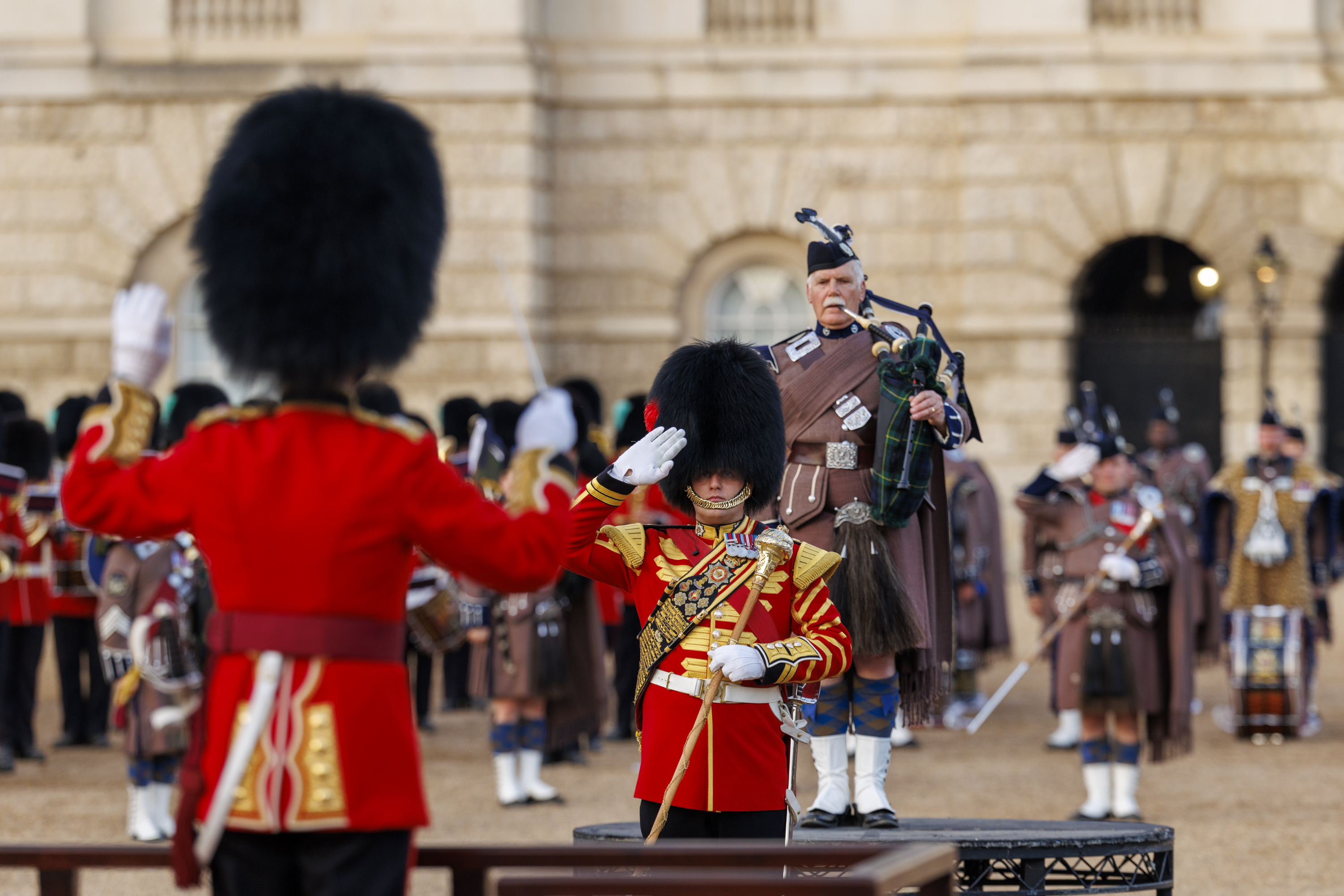 A person in a ceremonial red uniform with elaborate gold details salutes next to a guard. A bagpiper stands in the background wearing traditional Scottish attire, with other similarly dressed individuals visible. The setting appears formal, with historical architecture in the background.