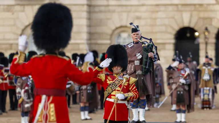 A person in a ceremonial red uniform with elaborate gold details salutes next to a guard. A bagpiper stands in the background wearing traditional Scottish attire, with other similarly dressed individuals visible. The setting appears formal, with historical architecture in the background.