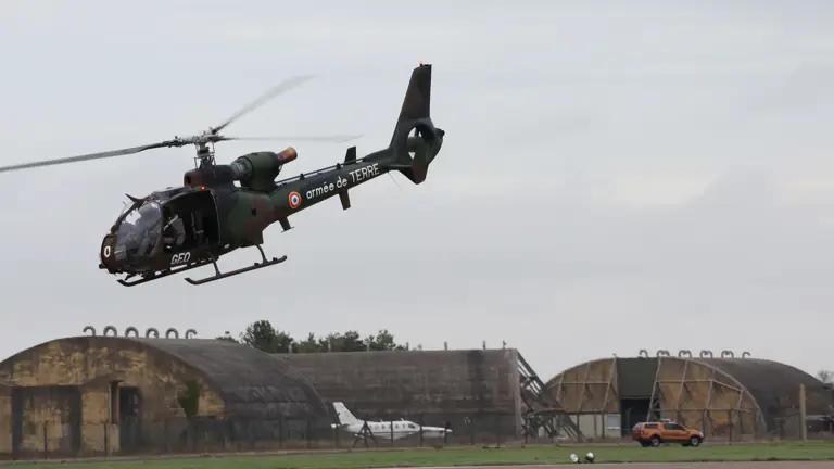 French Army light helicopter in flight above a military airfield with hangars and vehicles below.