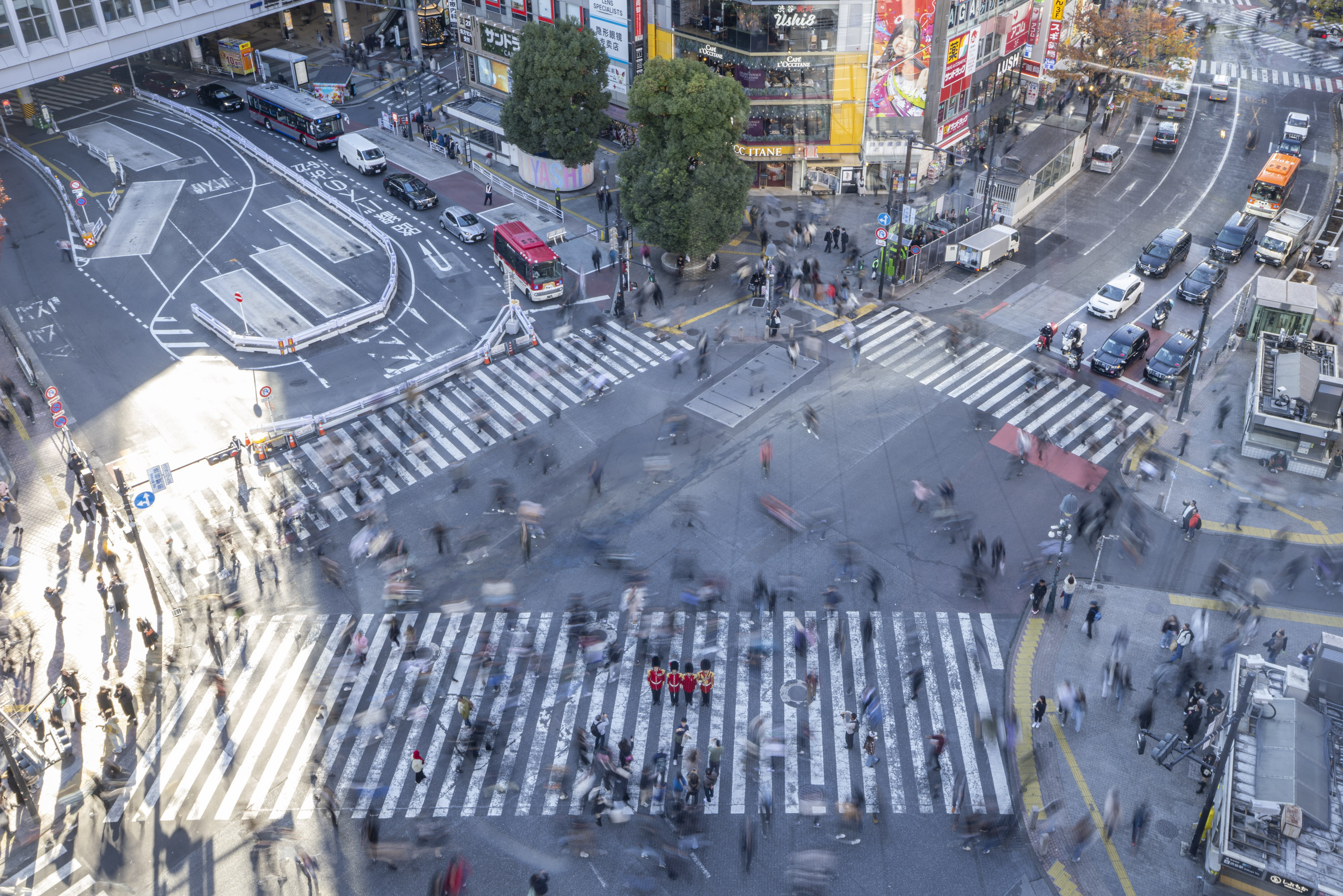 A bird's eye view shot of four soldiers in red tunics and bearskin hats on a crossing in Tokyo, pedestrians are a blur around them.