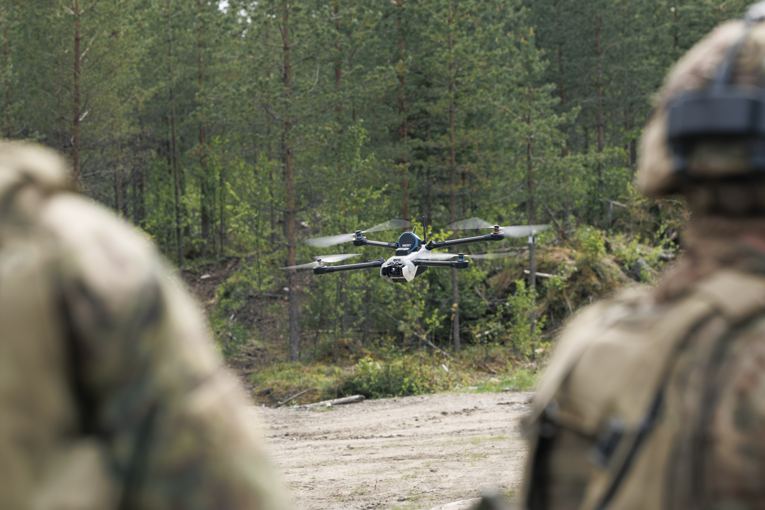 A drone hovers in a forested area, with blurred figures in camouflage gear in the foreground. Trees and greenery surround the scene.
