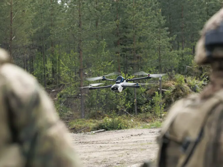 A drone hovers in a forested area, with blurred figures in camouflage gear in the foreground. Trees and greenery surround the scene.