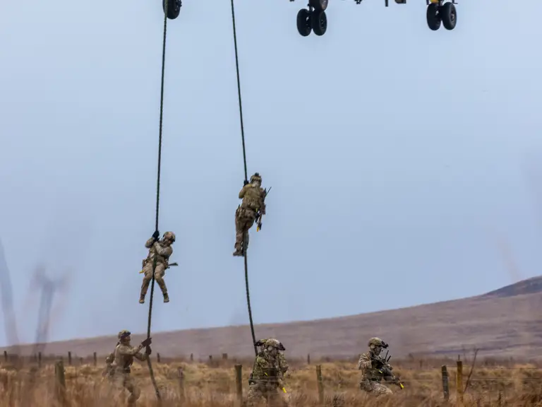 Military personnel rappelling from a hovering Chinook helicopter over a grassy field during a training exercise.