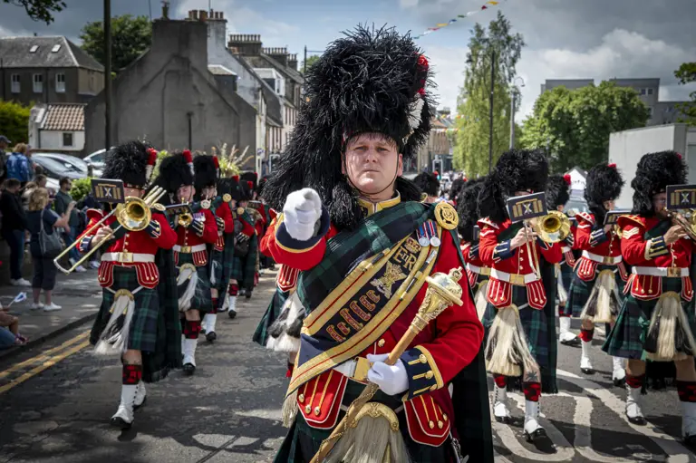 A soldier wears a red ceremonial tunic, sash and a black feather bonnet. He is leading a parade of musicians wearing red tunics and kilts.