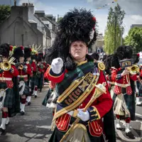 A soldier wears a red ceremonial tunic, sash and a black feather bonnet. He is leading a parade of musicians wearing red tunics and kilts.