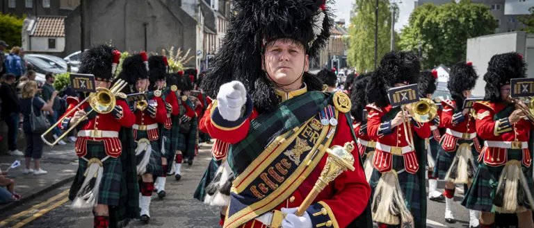 A soldier wears a red ceremonial tunic, sash and a black feather bonnet. He is leading a parade of musicians wearing red tunics and kilts.