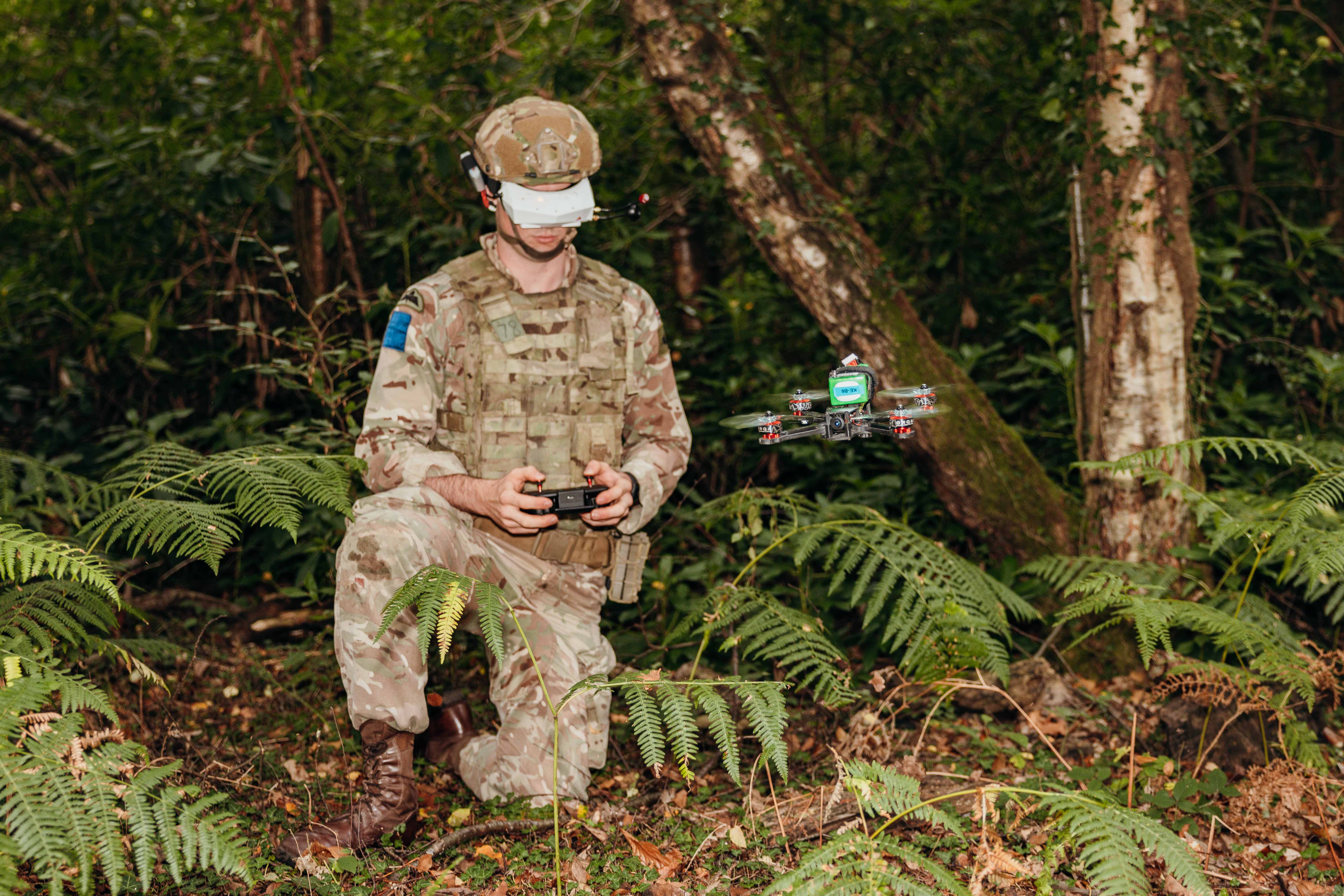 Soldier in camouflage uniform kneeling in a forest while controlling a drone with a remote device.