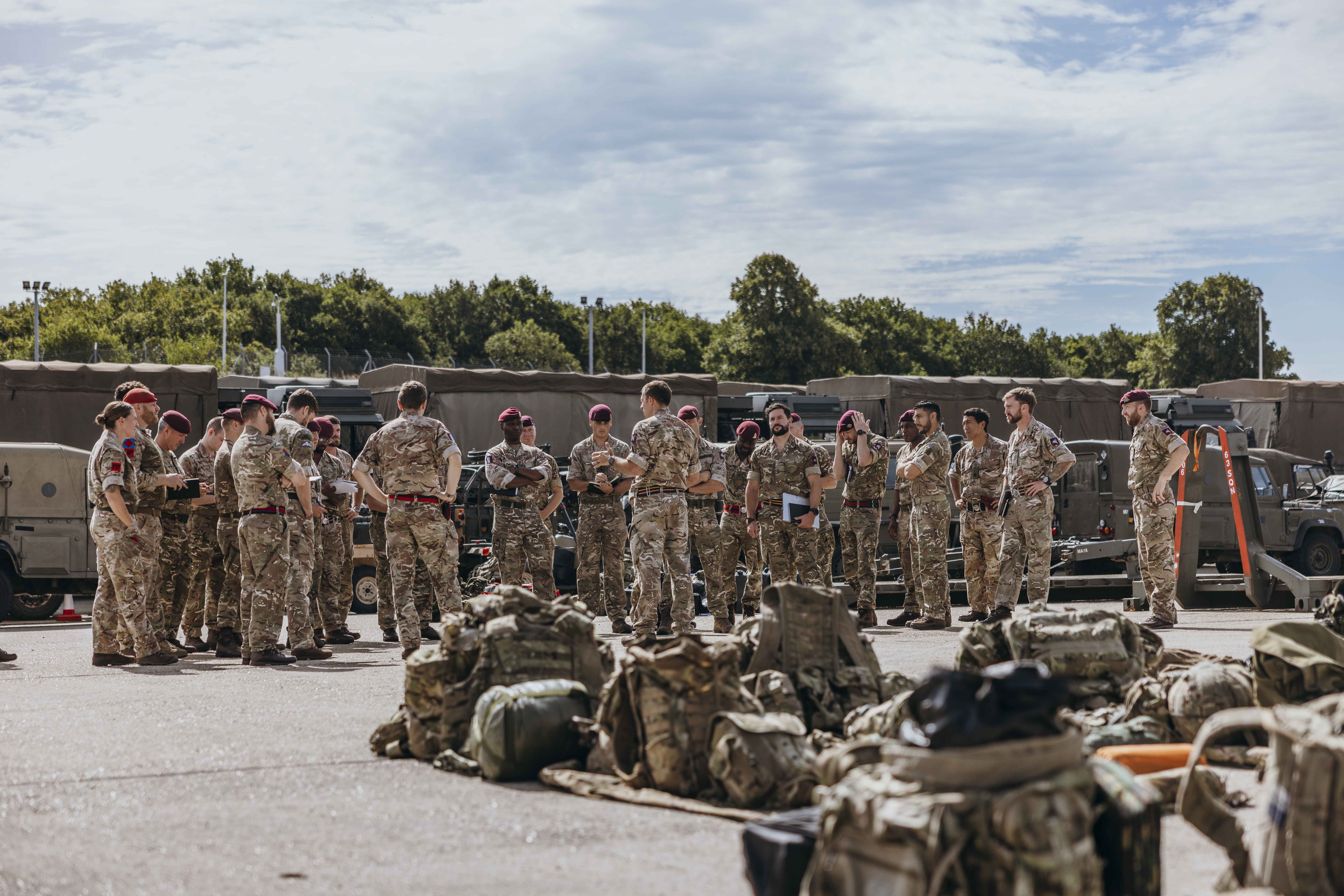 Group of soldiers in camouflage uniforms standing in a circle outdoors near military vehicles and equipment on a sunny day.