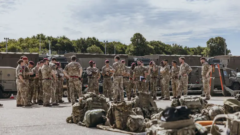 Group of soldiers in camouflage uniforms standing in a circle outdoors near military vehicles and equipment on a sunny day.