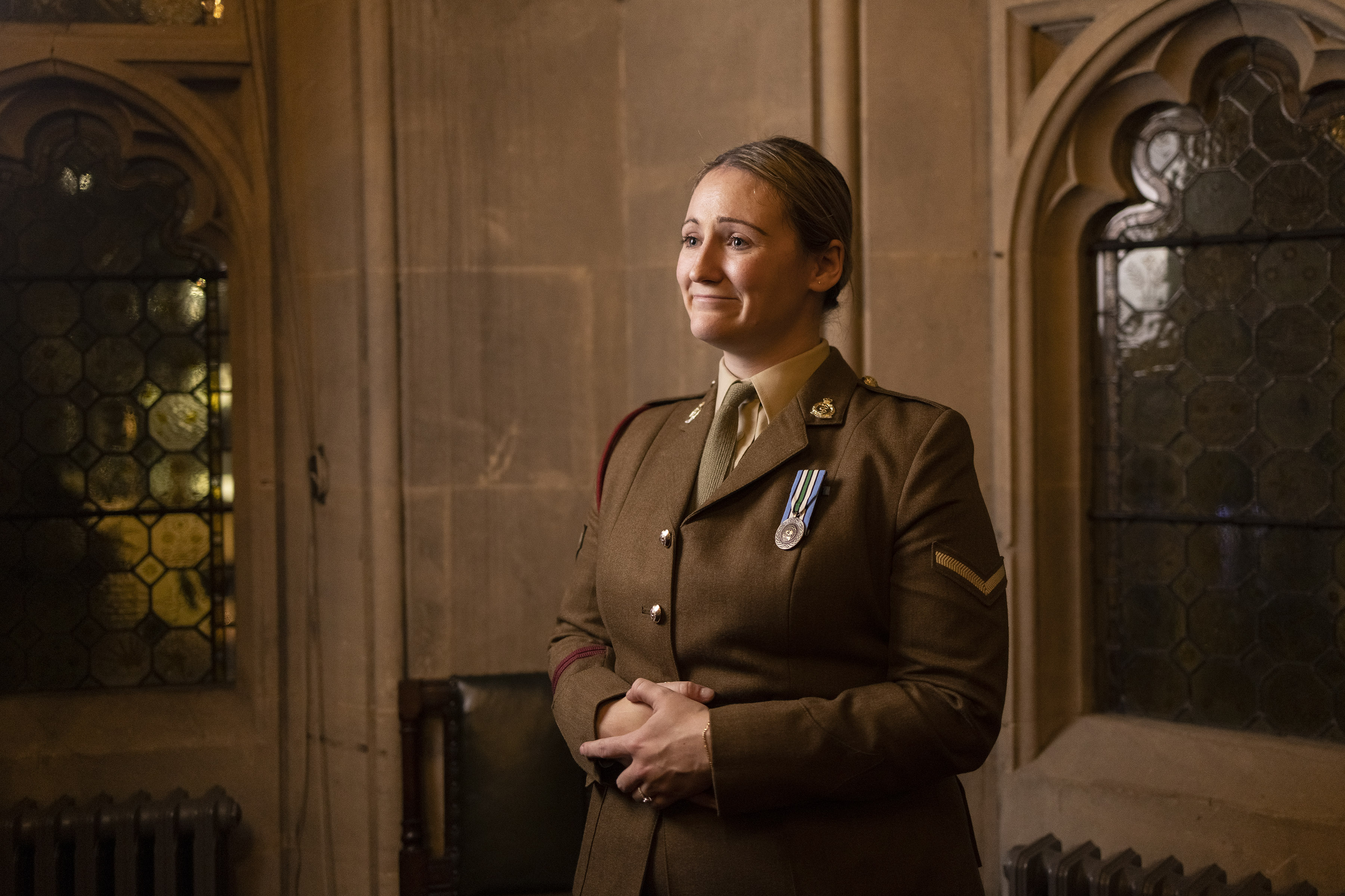 A woman in a military uniform stands in a dimly lit, ornate room. She wears medals on her chest and has a proud, reflective expression.
