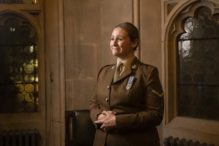 A woman in a military uniform stands in a dimly lit, ornate room. She wears medals on her chest and has a proud, reflective expression.