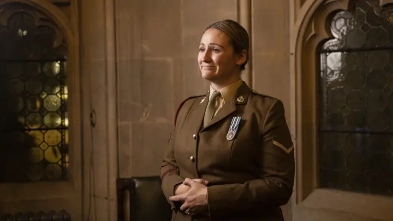 A woman in a military uniform stands in a dimly lit, ornate room. She wears medals on her chest and has a proud, reflective expression.