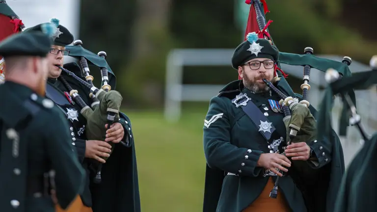 A bagpiper in a green and orange ceremonial uniform playing a bagpipe.