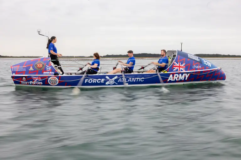 Four rowers in blue uniforms rowing a decorated boat labeled 'Force Atlantic Army' on calm water under a cloudy sky.