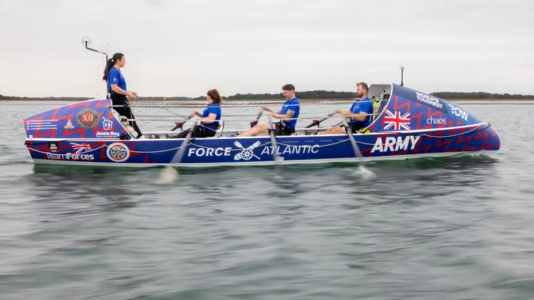 Four rowers in blue uniforms rowing a decorated boat labeled 'Force Atlantic Army' on calm water under a cloudy sky.