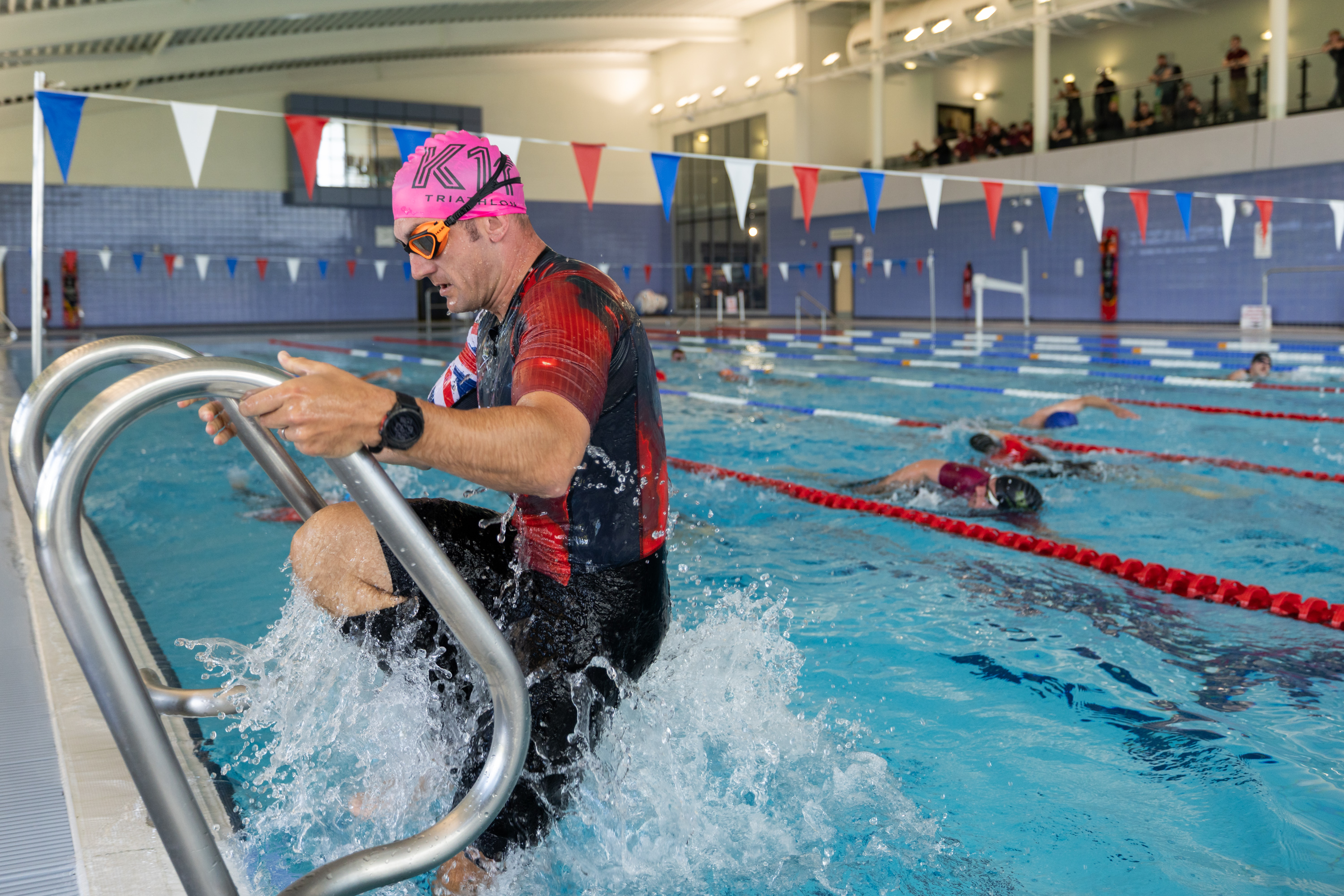 A person wearing a pink swim cap and goggles is exiting a swimming pool using a ladder. The pool is indoors, with multiple lanes and swimmers in the background. The setting appears to be a public or competition swimming facility.