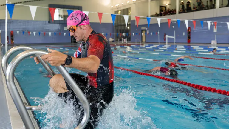 A person wearing a pink swim cap and goggles is exiting a swimming pool using a ladder. The pool is indoors, with multiple lanes and swimmers in the background. The setting appears to be a public or competition swimming facility.