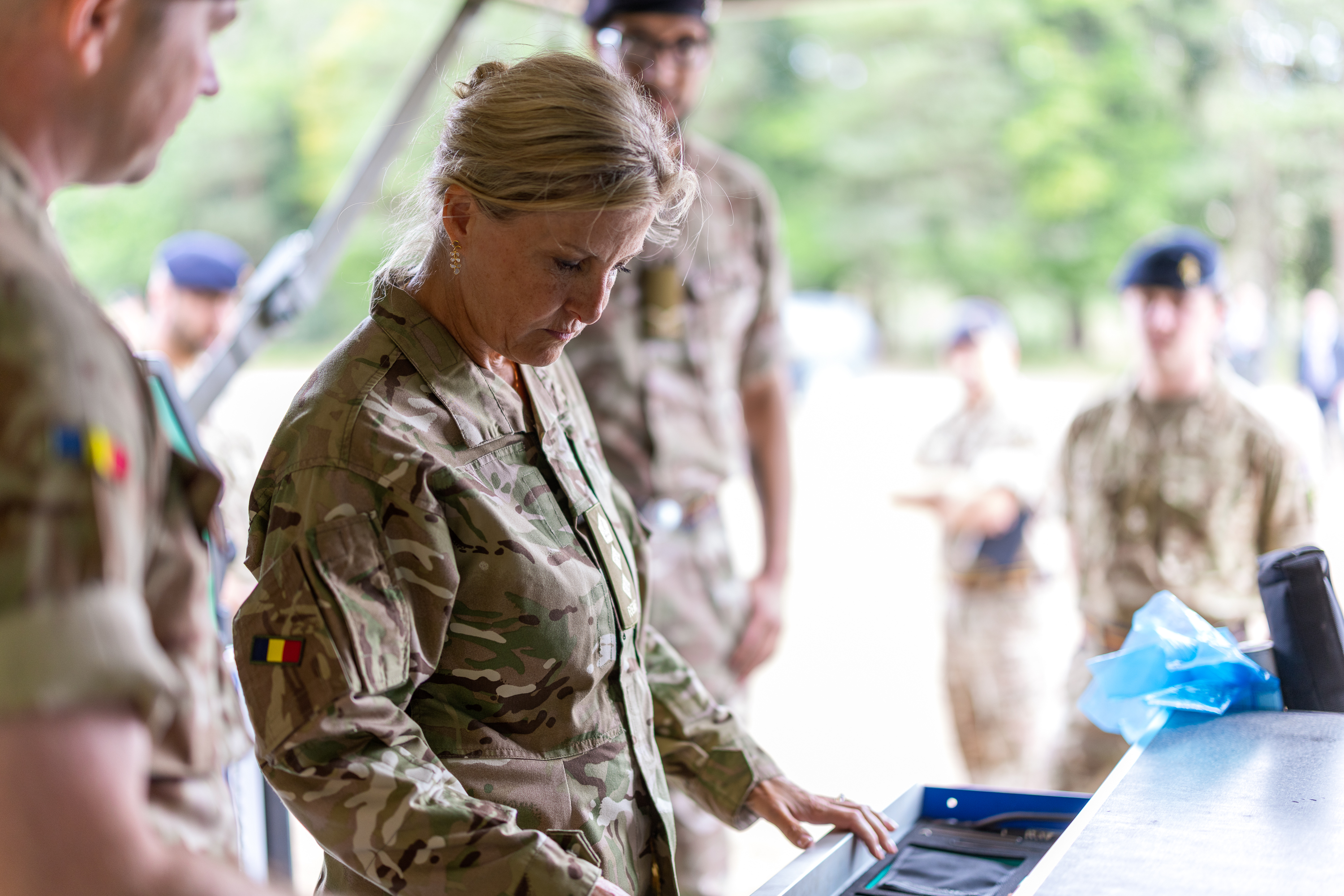 The Duchess of Edinburgh is seen wearing trousers and a camouflage print jacket stood outside with two soldiers in camouflage uniform on stood either side of her.