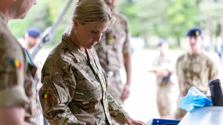 The Duchess of Edinburgh is seen wearing trousers and a camouflage print jacket stood outside with two soldiers in camouflage uniform on stood either side of her.
