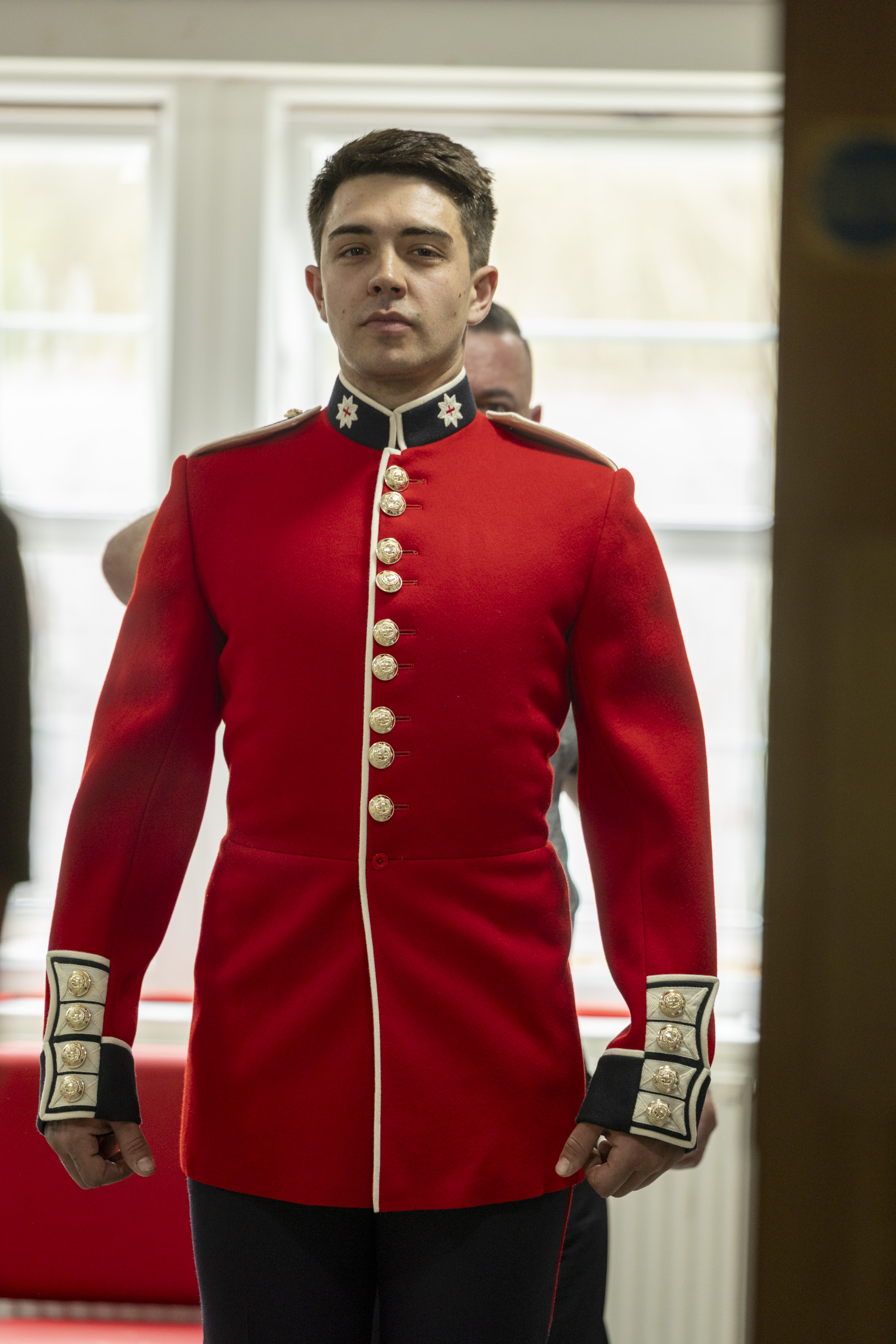 This image shows someone buttoning a red tunic as the ceremonial dress of the Household Division. The jacket features coldstream guards cap badge imprinted on shiny, metallic buttons going down the cuff of the tunic, and the front of the tunic.
