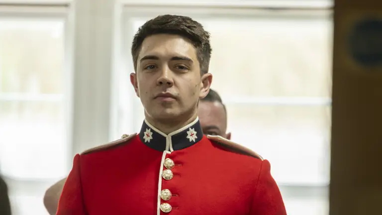 This image shows someone buttoning a red tunic as the ceremonial dress of the Household Division. The jacket features coldstream guards cap badge imprinted on shiny, metallic buttons going down the cuff of the tunic, and the front of the tunic.