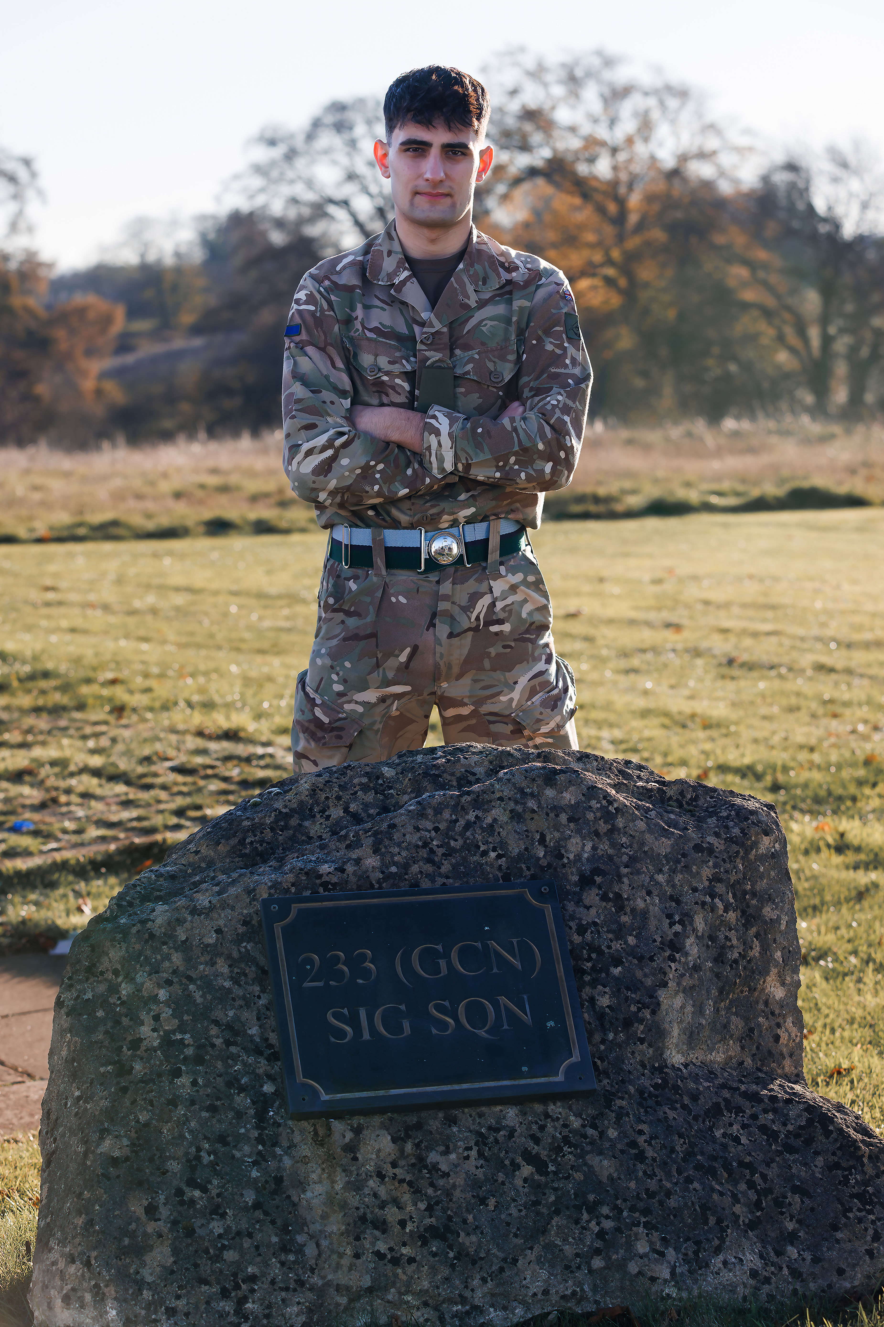 A person in military camouflage stands confidently with crossed arms behind a plaque on a boulder reading 
