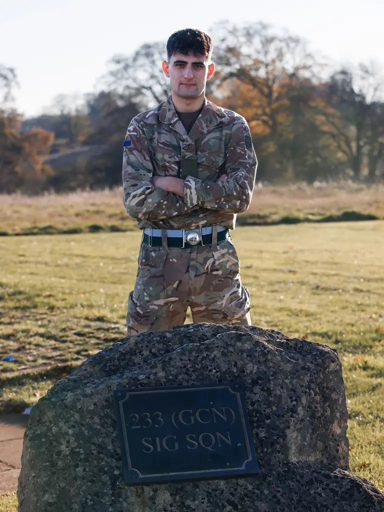 A person in military camouflage stands confidently with crossed arms behind a plaque on a boulder reading
