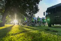 Runners in bright gear sprint across a sunlit park under large trees. The setting sun creates dramatic shadows; a truck is parked nearby. Energetic atmosphere.